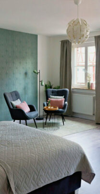 MainZauber Apartments früher MainTerrasse Miltenberg am Main - Modernes Schlafzimmer in einer luxuriösen Ferienwohnung mit blauer Akzentwand, großen Fenstern und stilvollen Sitzgelegenheiten.