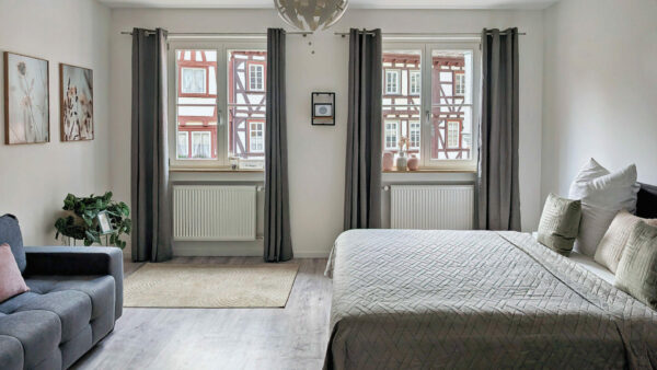 MainZauber Apartments früher MainTerrasse Miltenberg am Main - Modernes, helles Schlafzimmer in einer luxuriösen Ferienwohnung in der Altstadt mit großem Bett, Sofa und Blick auf Fachwerkhäuser.