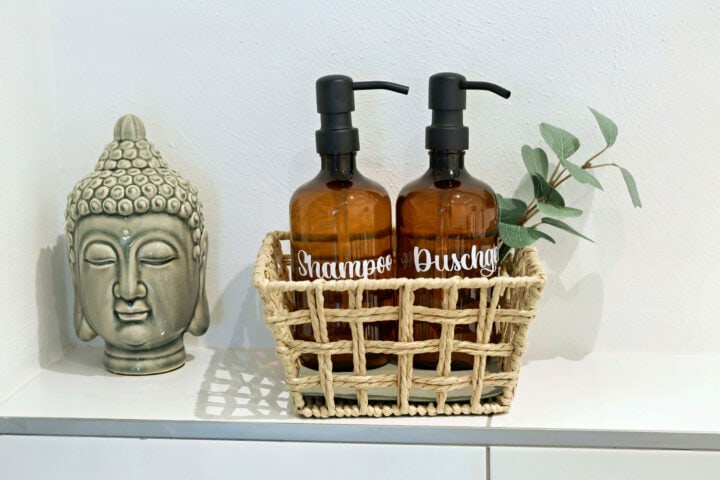 MainZauber Apartments früher MainTerrasse Miltenberg am Main - Zwei bernsteinfarbene Spender für Shampoo und Duschgel im Korb, neben Buddha und Eukalyptus - Luxus-Apartment Flair.