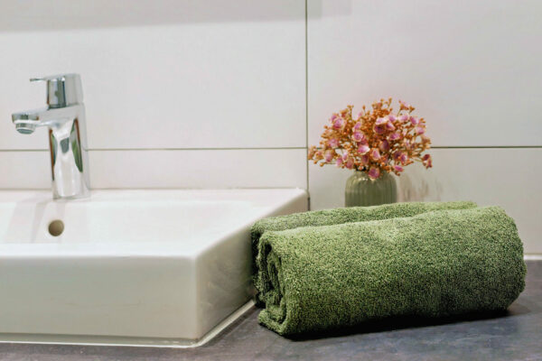 MainZauber Apartments früher MainTerrasse Miltenberg am Main - Grünes Handtuch auf Badablage, neben weißem Waschbecken mit Chromarmatur, Deko-Blumen - luxuriöse Ferienwohnung Atmosphäre.