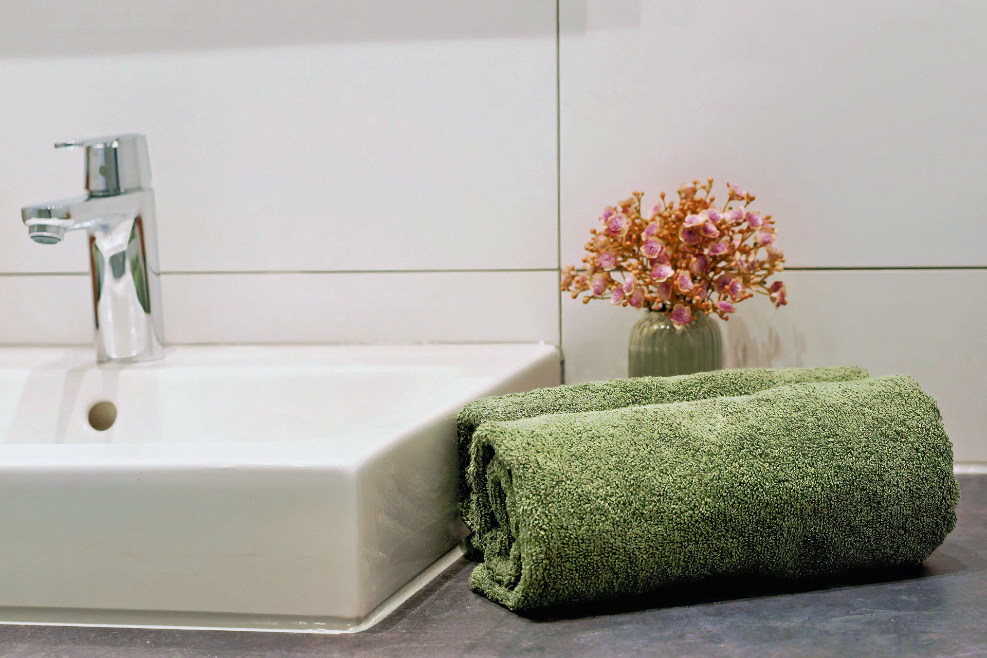 MainZauber Apartments früher MainTerrasse Miltenberg am Main - Grünes Handtuch auf Badablage, neben weißem Waschbecken mit Chromarmatur, Deko-Blumen - luxuriöse Ferienwohnung Atmosphäre.
