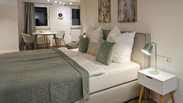 MainZauber Apartments früher MainTerrasse Miltenberg am Main - Entdecken Sie die perfekte Ferienwohnung für Ihren nächsten Kurzurlaub. Diese luxuriöse Unterkunft verfügt über ein gemütliches Schlafzimmer mit einem ordentlich gemachten Bett, das mit eleganter grün-weißer Bettwäsche ausgestattet ist. Genießen Sie den Komfort von zwei Nachttischen mit Lampen, die für einen angenehmen Aufenthalt sorgen. Genießen Sie Ihre Mahlzeiten am kleinen Esstisch mit Stühlen in der Nähe des Fensters, das Ihnen beim Essen eine einladende Aussicht bietet. Die neutrale Einrichtung der Wohnung, ergänzt durch geschmackvolle Wandkunst und sanfte Beleuchtung, schafft eine warme und einladende Atmosphäre, die ideal zum Entspannen während Ihres Urlaubs ist.